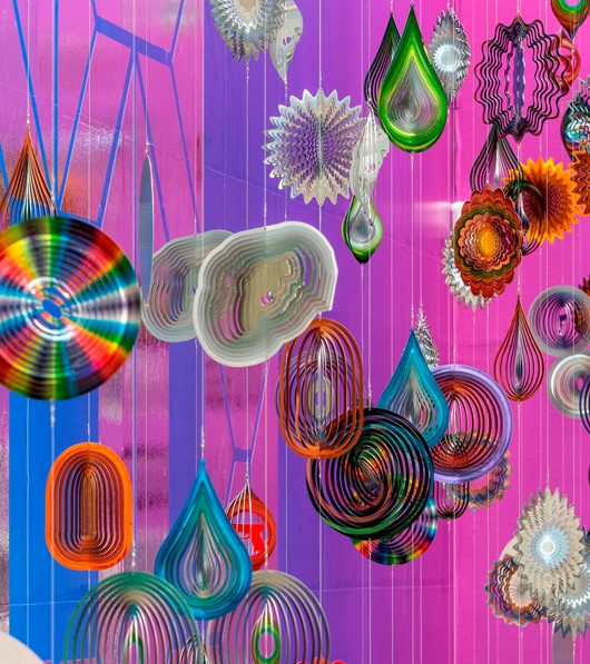 A collection of small metal sculptures hang on wires against a dazzling pink and purple backdrop