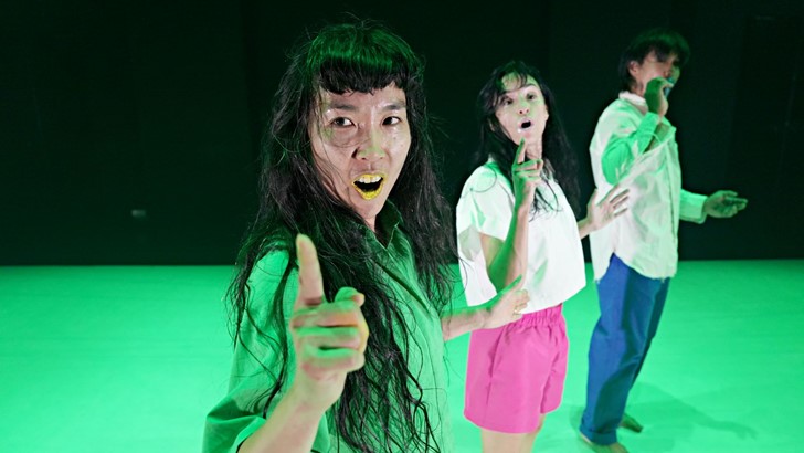 Three dancers stand on a bright green stage with a dark backdrop, they look towards the photographer