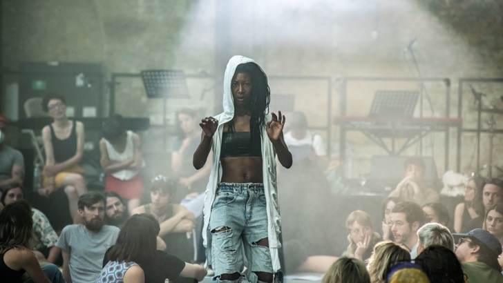 A performer stands, hands raised dramatically, watched by an audience seated casually in a gallery.