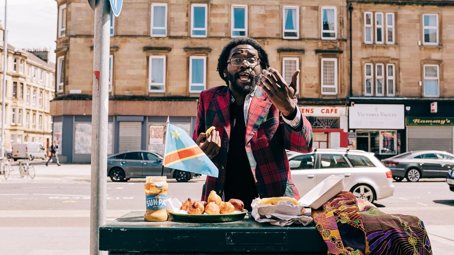 A performer raises his hand expressively to the viewer. He stands behind a table sitting in an urban street. It bears a plate of fried food and a Congolese flag.
