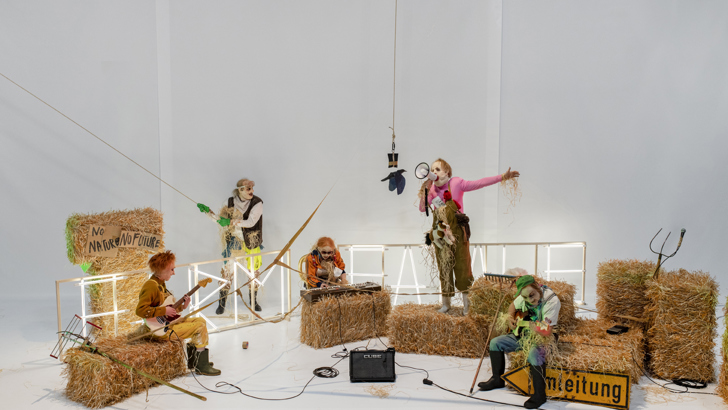 A group of performers in unsettling masks share a brightly lit stage with bales of hay