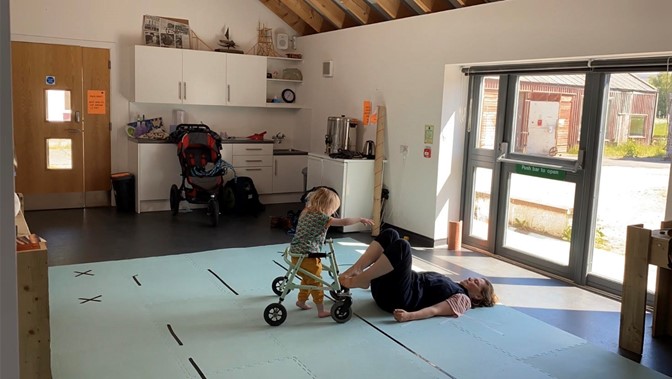 In a studio, a woman lies on the floor, her feet on a walking frame which is being pushed by a young child