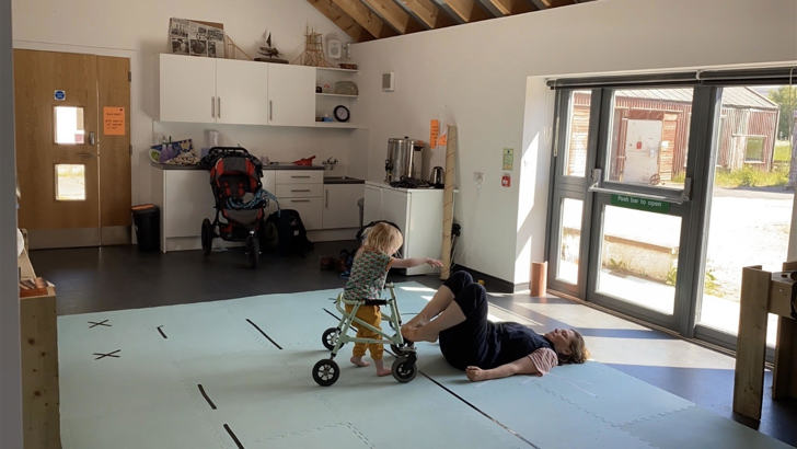 In a studio, a woman lies on the floor, her feet on a walking frame which is being pushed by a young child