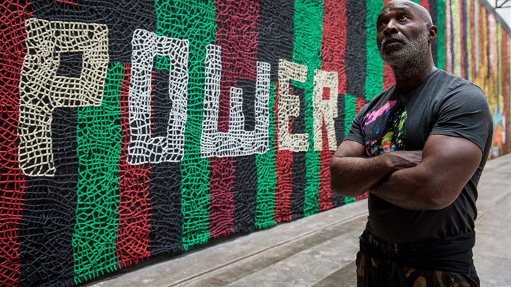 The artist Nick Cave poses, arms folded, against his work; a large wall mural which depicts the word 'power'