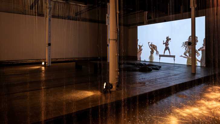 A gallery space displaying a video of figures in straw masks and traditional Cambodian costume, behind an artificial wall of rain