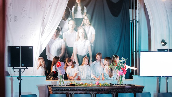 School children pose in front of a scaled up photo of themselves, partially hidden by a stage curtain