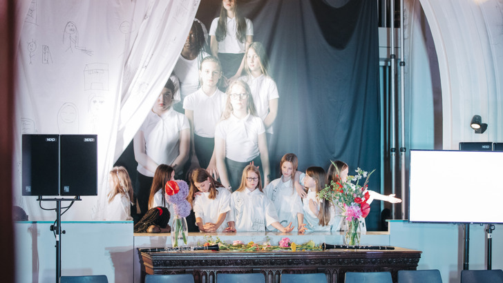 School children pose in front of a scaled up photo of themselves, partially hidden by a stage curtain