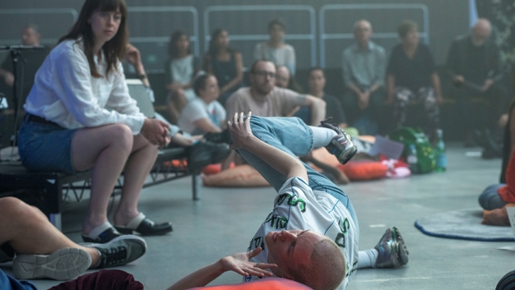 People congregate in a large gallery, some seated on chairs and on the floor. A figure in the foreground sprawls on the floor. 