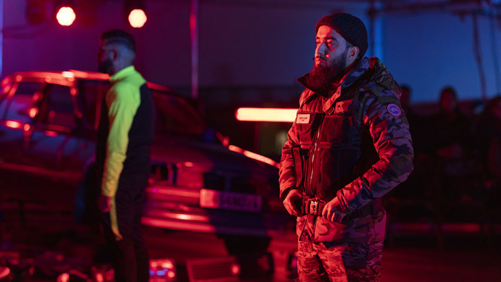 Two men, one slightly out of focus, stand in front of a car on a dramatically lit set