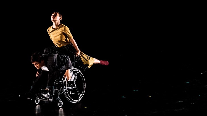 A duo, one in a wheelchair, dance together on a dark stage