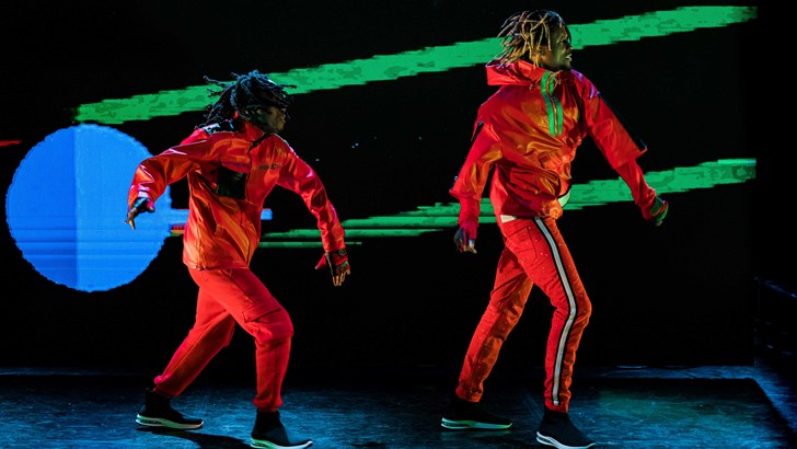 Two figures in high vis costume strike the same pose against a brightly coloured abstract projection