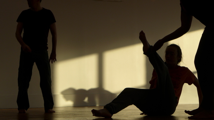 Three figures silhouetted in a studio space, one seated on the floor