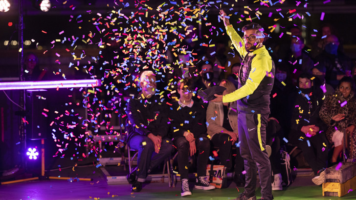 A man in a tracksuit looks on as confetti flutters around him. There is a seated audience in the background.