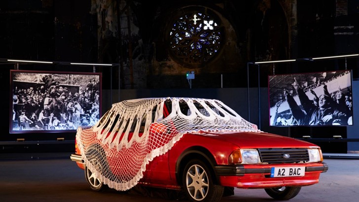 A red car draped in a vast doily sits in front of two large black and white tapestries