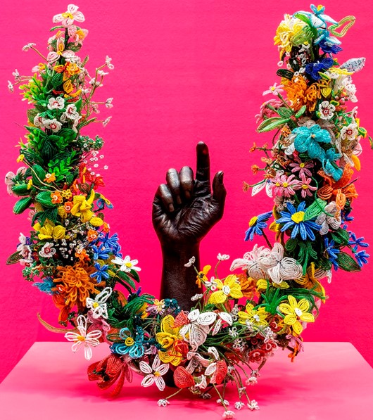A black sculpture of a hand, one finger raised to the sky, sits behind a horseshoe garland of flowers