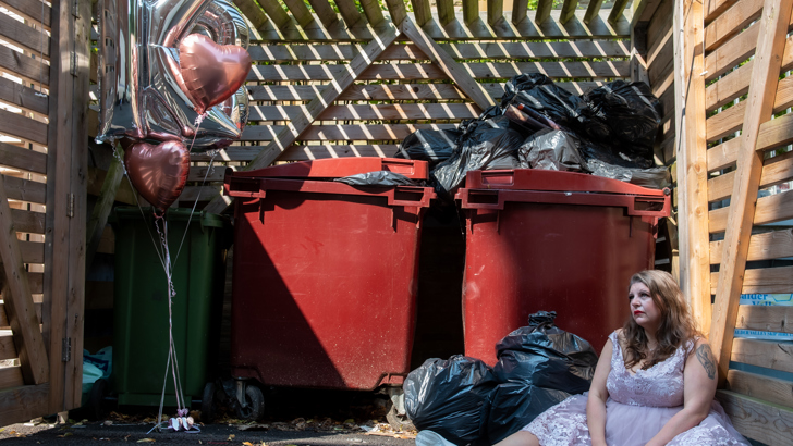 A woman sits in a bin store, flanked by heart shaped balloons. She wears a party dress and a non-plussed expression 