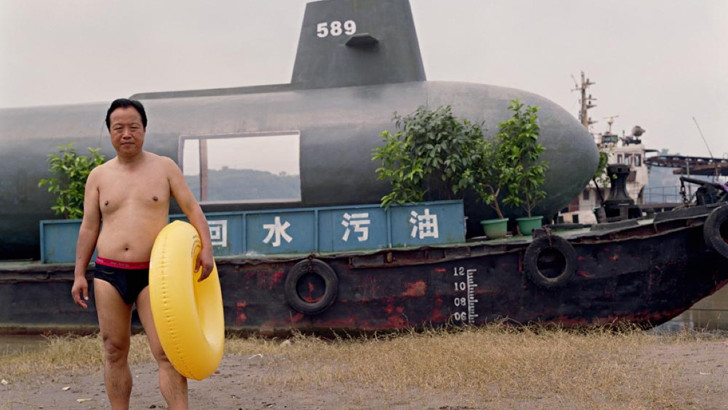 A man in swimming trunks, holding a rubber ring, poses incongruously in front of grounded submarine