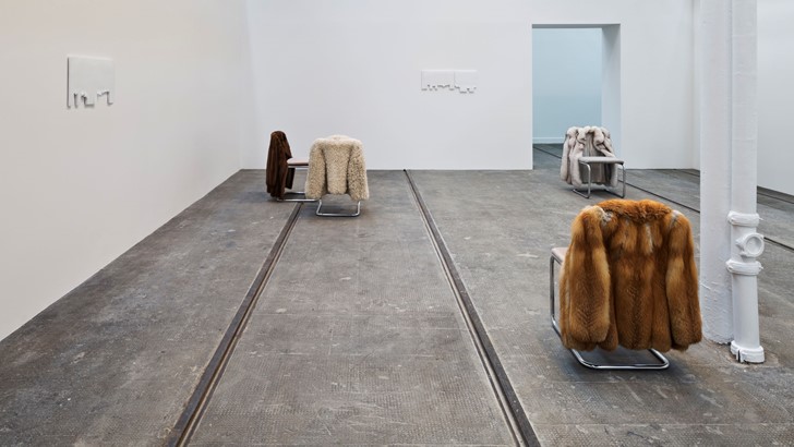 A white washed gallery sparsely populated by fur coats hung over chairs