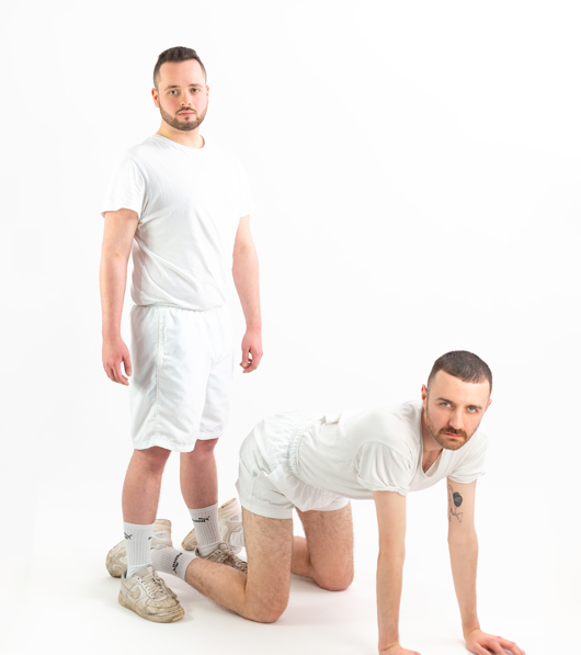 Two men in matching matching sports outfits pose together, one kneeling in front of the other