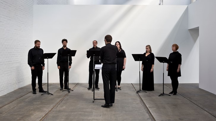 Six singers in dark clothes stand behind music stands. A conductor faces them, his back to the viewer