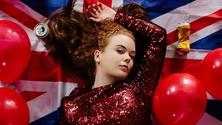 A performer in a sequined dress lies surrounded by balloons, beer cans and a union jack
