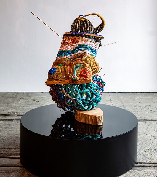 A colourful woven abstract sculpture sits on a black plinth