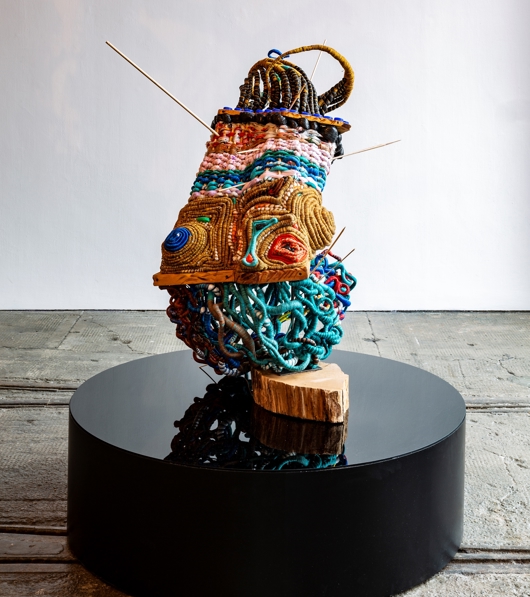 A colourful woven abstract sculpture sits on a black plinth