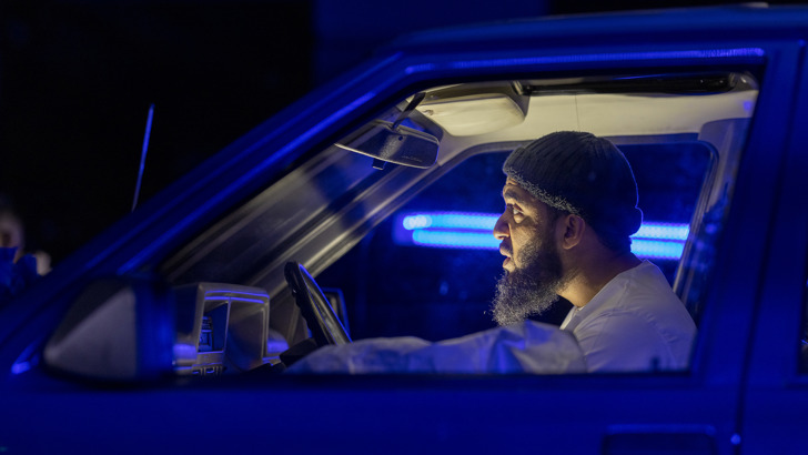 A man sits in a car on a dramatically lit set, his serious face in profile