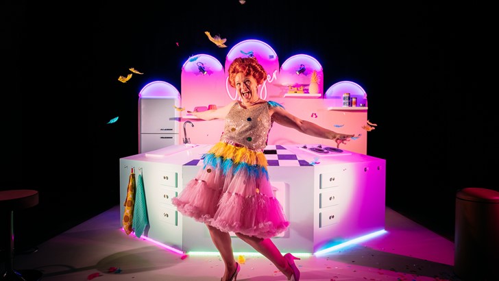 A performer in a multicoloured dress poses in front of a kitsch kitchen stage set