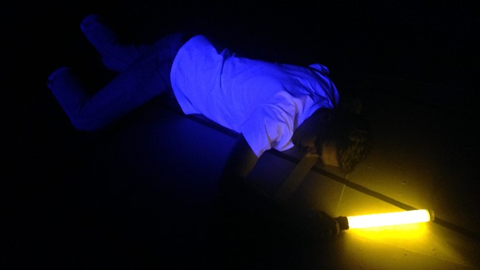 A performer lies in semi-darkness holding a light tube