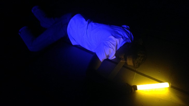 A performer lies in semi-darkness holding a light tube