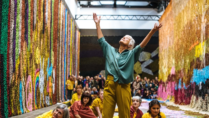 A woman poses with arms raised, surrounded by other performers. In the distance, a crowd of spectators watch them.