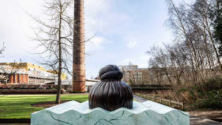 A sculpture depicts the top of a persons head emerging from a marble-effect surface. The sculpture is in a garden, surrounded by greenery