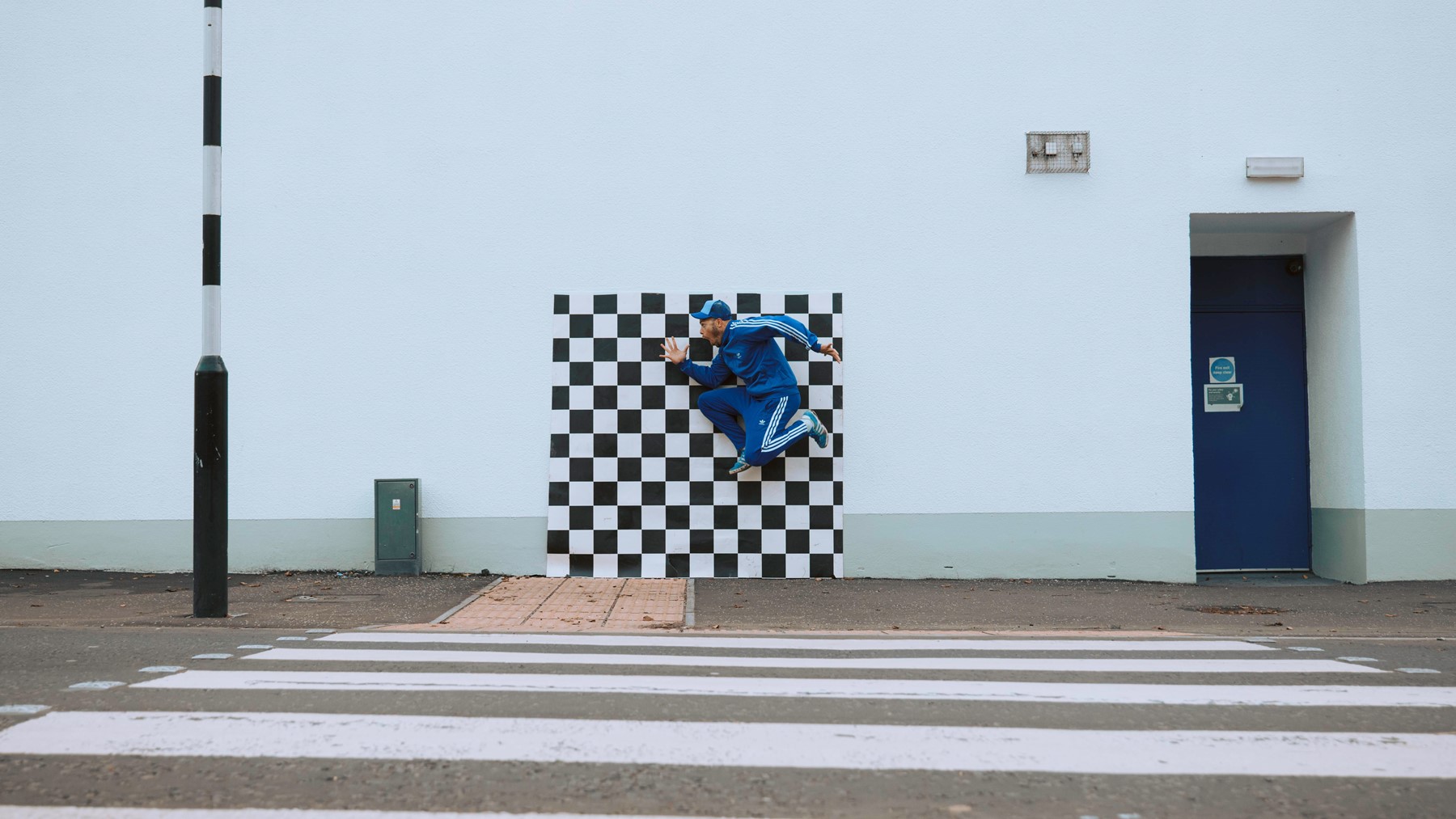 A performer in a tracksuit jumps high against a checkerboard vinyl, which sits in front of an urban scene