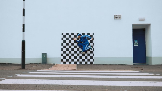 A performer in a tracksuit jumps high against a checkerboard vinyl, which sits in front of an urban scene