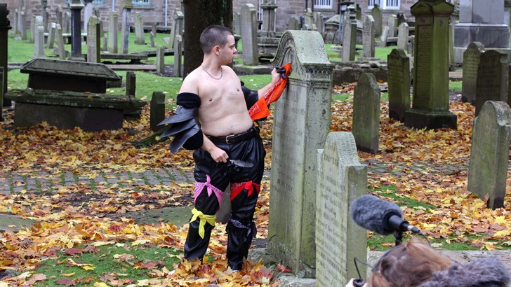A topless figure looks intent as they wipe a gravestone with a red scarf