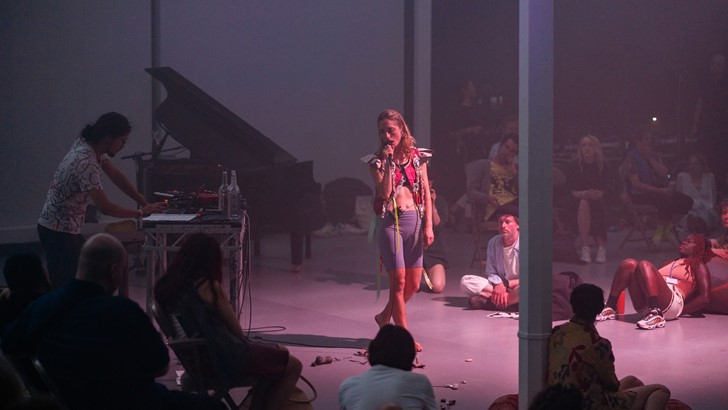 A performer speaks into a microphone, watched by an audience seated casually in a gallery.