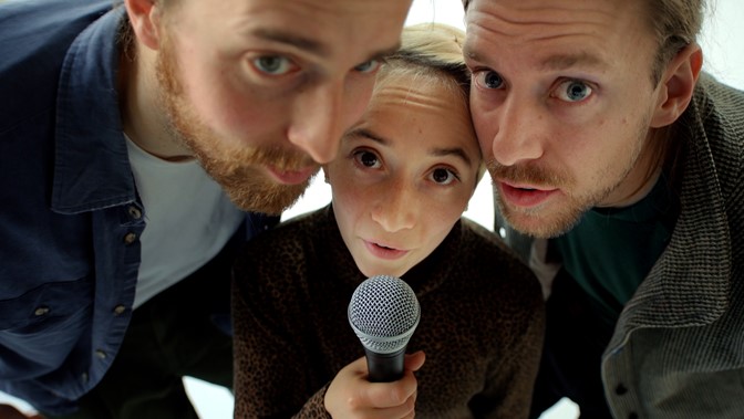 Three figures huddle round a microphone, gazing at the viewer quizzically