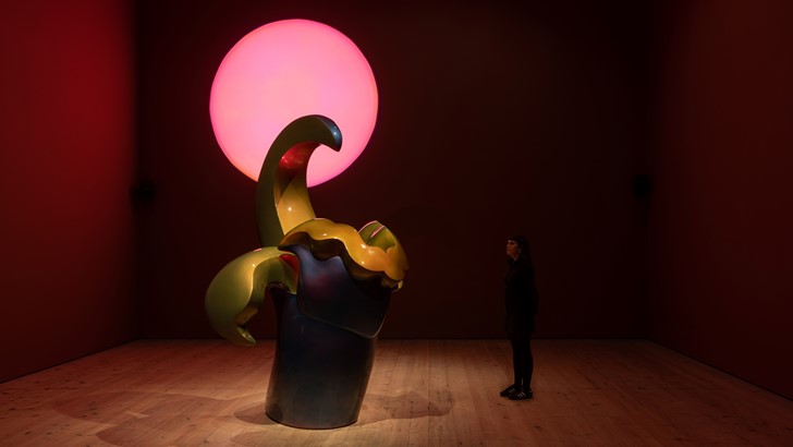 In a darkened gallery, a large abstract sculpture dwarves a lone viewer. Behind it glows a pink sun-like orb