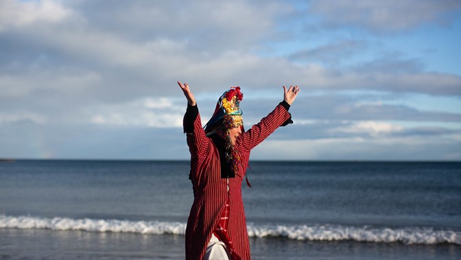 A woman in folk costume poses, arms aloft, in front of a seascape