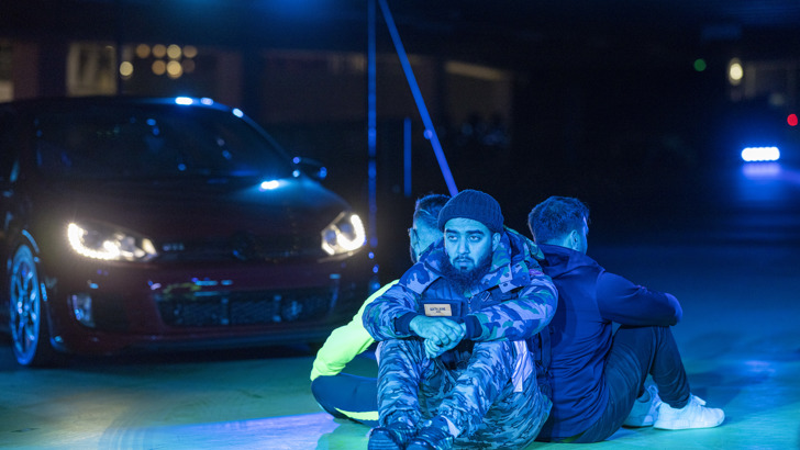 Three men sit back to back in front of a car on a dramatically lit set