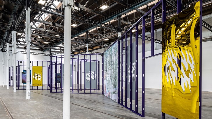 Metal sculptures and screens are displayed on a cage-like steel structure in a large gallery