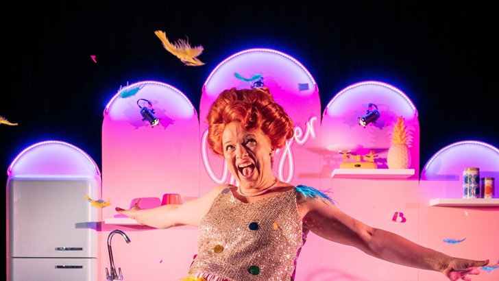A beehived performer poses exuberantly in front of a pink lit stage set