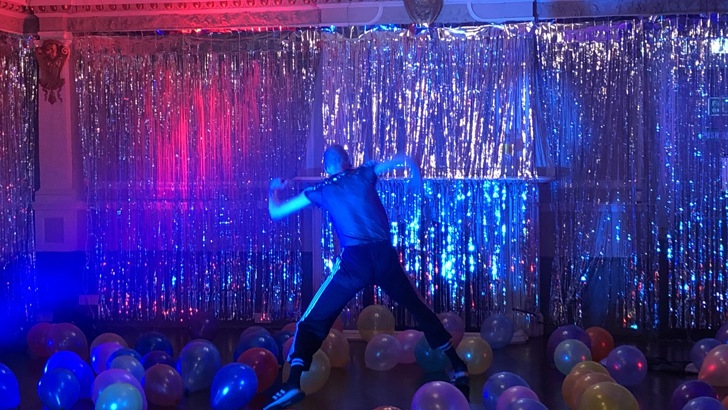 A figure dances against a shimmer curtain, the stage strewn with balloons