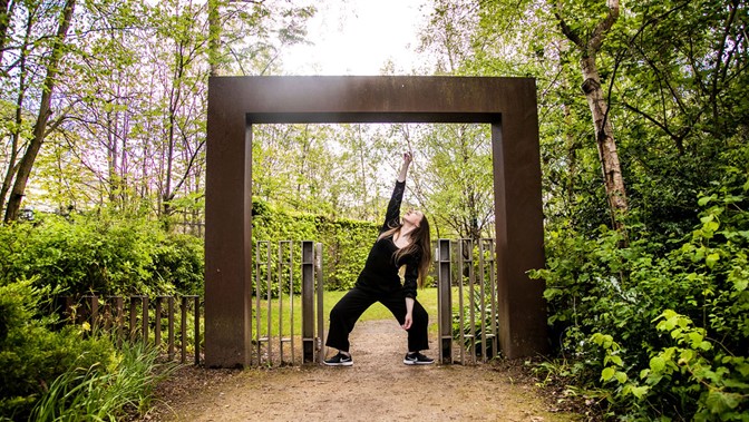 A performer poses in a woodland setting, framed by a modernist steel structure