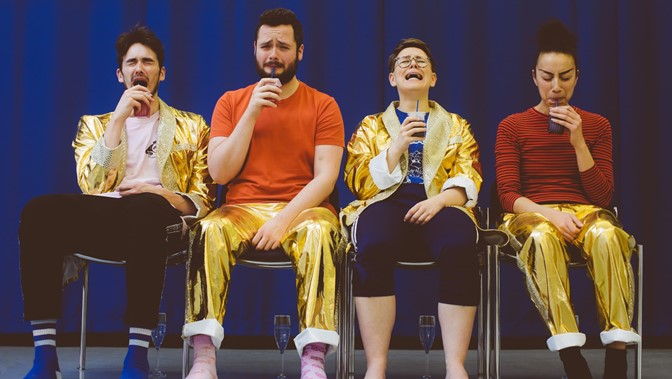 Four worried-looking performers are seated in a row, on stage