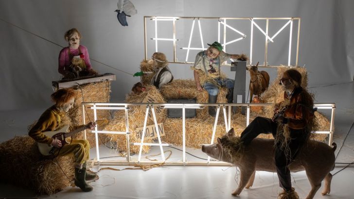 A group of performers in unsettling masks share a brightly lit stage with bales of hay and a model pig