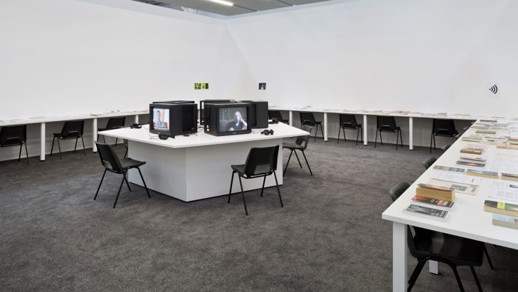 Desks covered in books line three walls of a gallery. The centre of the room holds more desks, on which sit televisions.