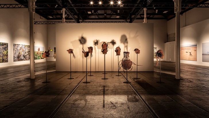 A gallery space displaying a collection of dramatically lit straw masks 